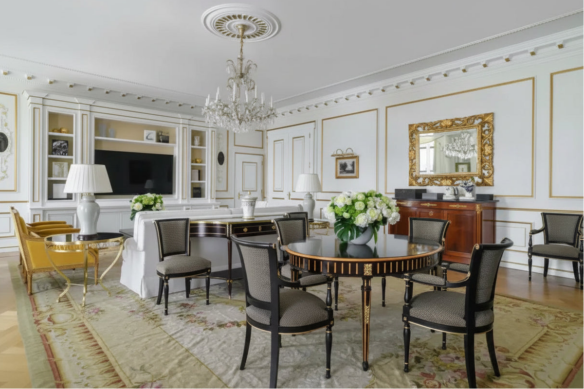 An elegant interior room featuring a round wooden dining table with upholstered chairs, a crystal chandelier, built-in shelving, a TV, and a gold-framed mirror on white paneled walls.