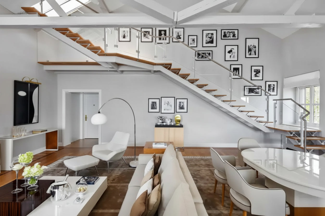 A modern open-plan living space with a staircase, dining table, sofa, chairs, coffee table, and framed black-and-white wall art. The room has white walls, exposed white ceiling beams, wood flooring, and large windows.