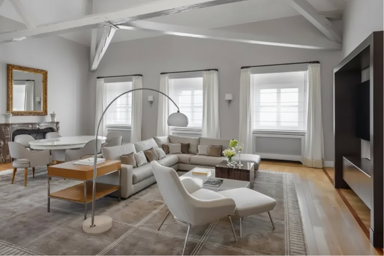 A modern living room featuring a light gray sectional sofa, a white lounge chair, a tall arched floor lamp, and a large patterned area rug. The room has high ceilings with exposed white beams, large windows with white curtains, and light wood floors.
