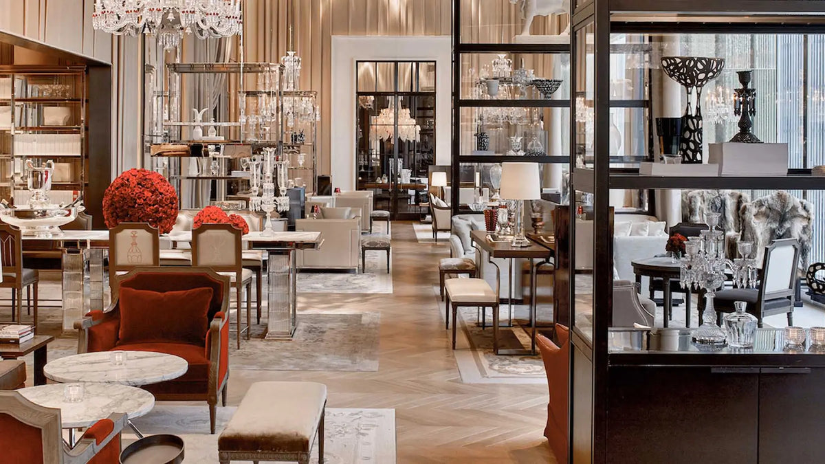 A luxury interior showroom with elegant dining tables, upholstered chairs, chandeliers, shelving, and decorative glass display cases in a warm beige, brown, and black color palette.