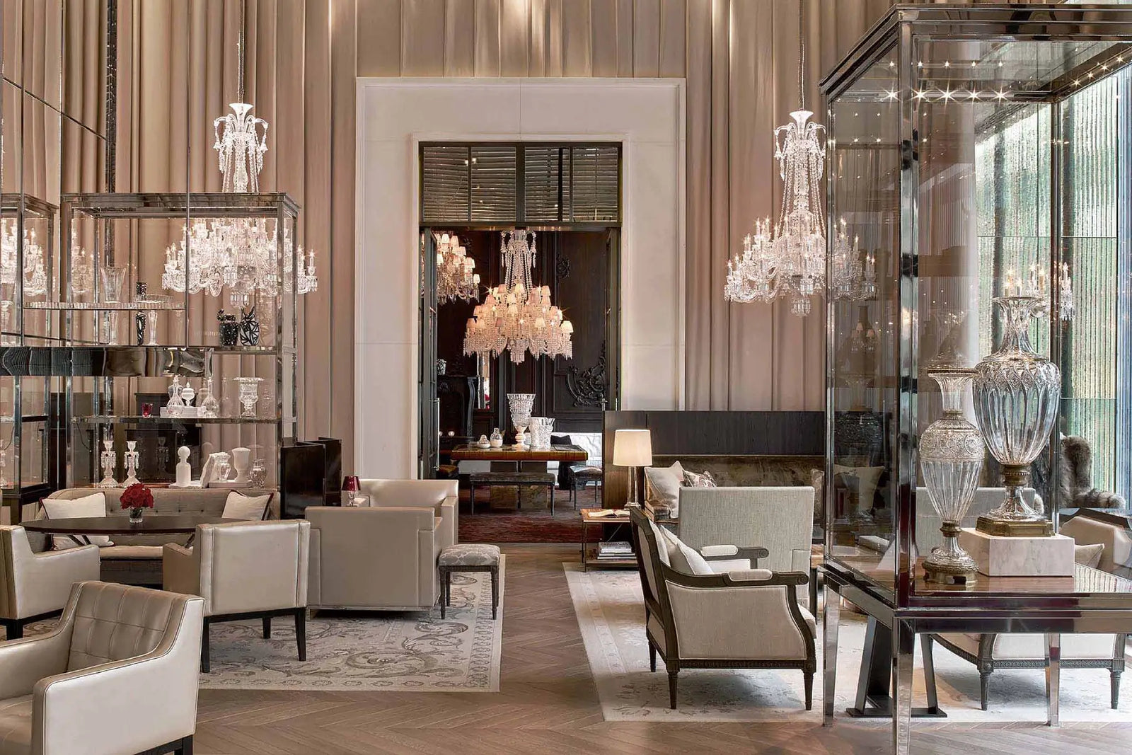 An elegant interior showroom or lounge with multiple crystal chandeliers, upholstered seating, glass display cases, and decorative vases in a luxurious beige and brown setting.
