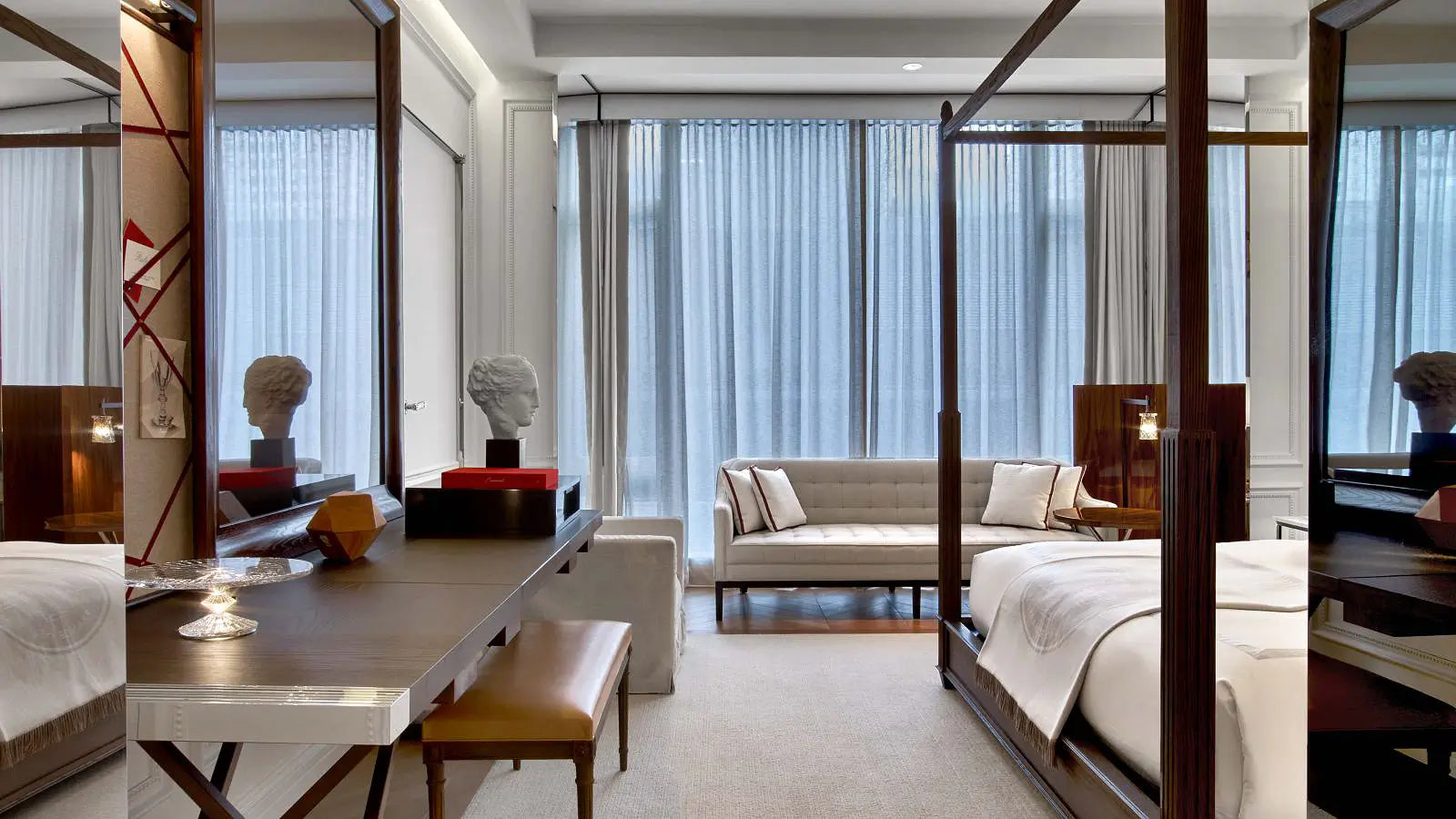 A luxury modern bedroom featuring a dark wood four-poster bed, a matching wooden vanity desk with a bench, a light beige sofa, and large sheer curtains in the background.