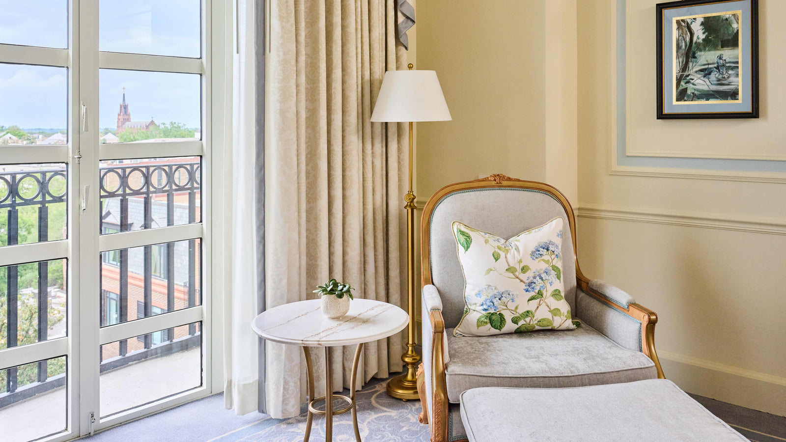 A styled interior corner featuring a cream upholstered armchair with a floral throw pillow, a small round marble side table, a gold floor lamp, and a window with beige curtains overlooking a balcony.