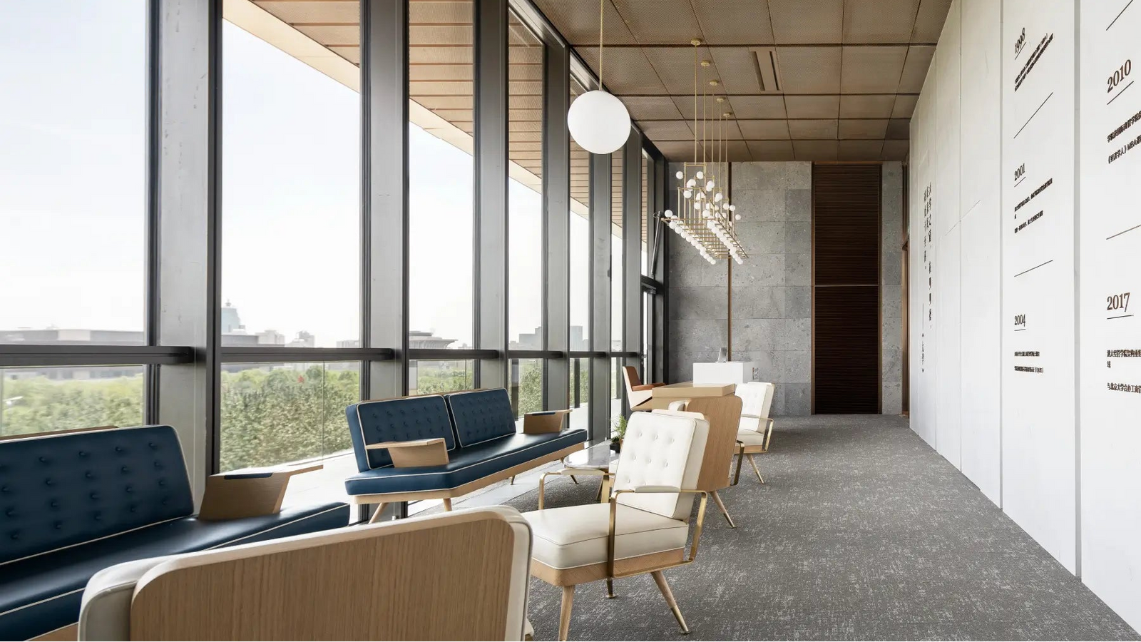 A modern office lounge area with large floor-to-ceiling windows, light wood ceiling panels, and seating including blue upholstered sofas, cream armchairs, and small tables. No people are present, and the space is shown as an interior environment rather than a single isolated product.