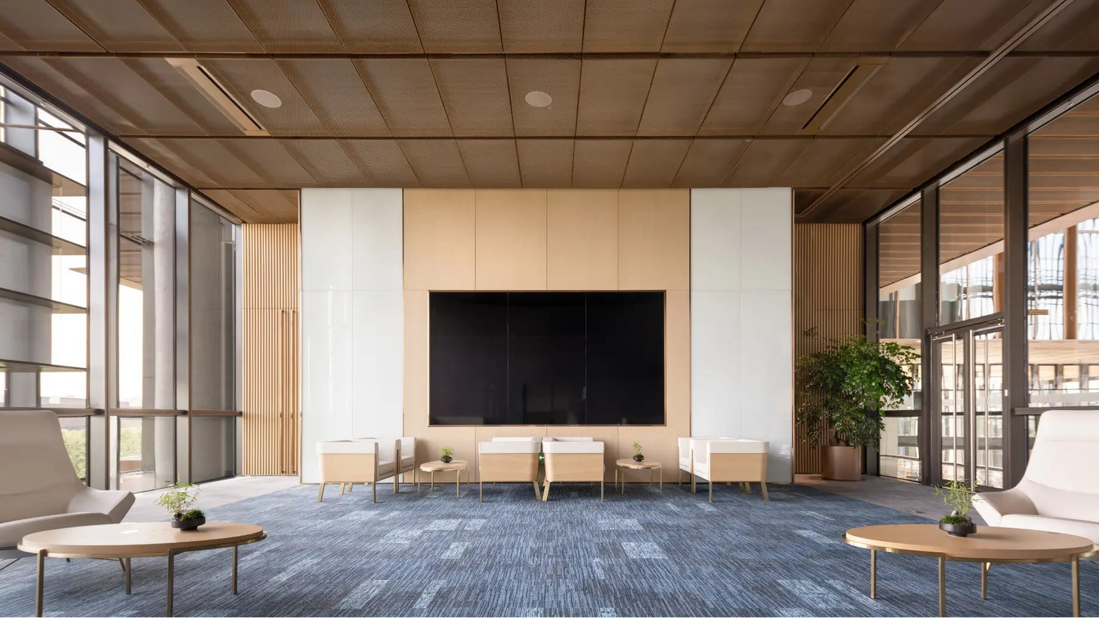 A modern, spacious interior lounge or meeting space with a large black wall-mounted screen centered on a light wood and white panel wall. The room has a blue-gray patterned carpet, a wood ceiling, large windows on both sides, and several minimalist chairs and round tables.