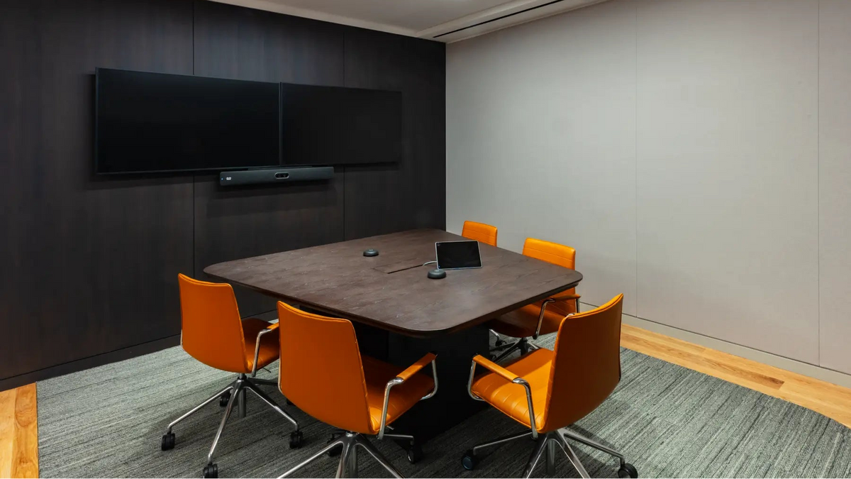 Modern conference room with a dark wood rectangular table, six orange swivel chairs, a large wall-mounted video conference screen, gray carpet, and light gray paneled walls.