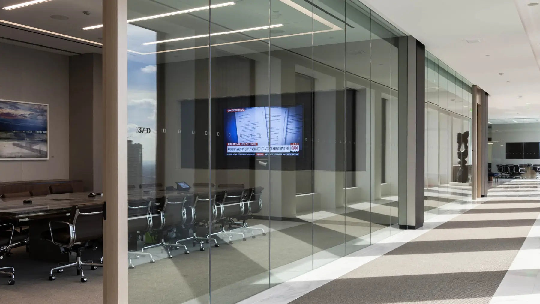 Modern glass-walled conference room with a large wall-mounted screen, meeting chairs, and an office hallway with striped flooring.