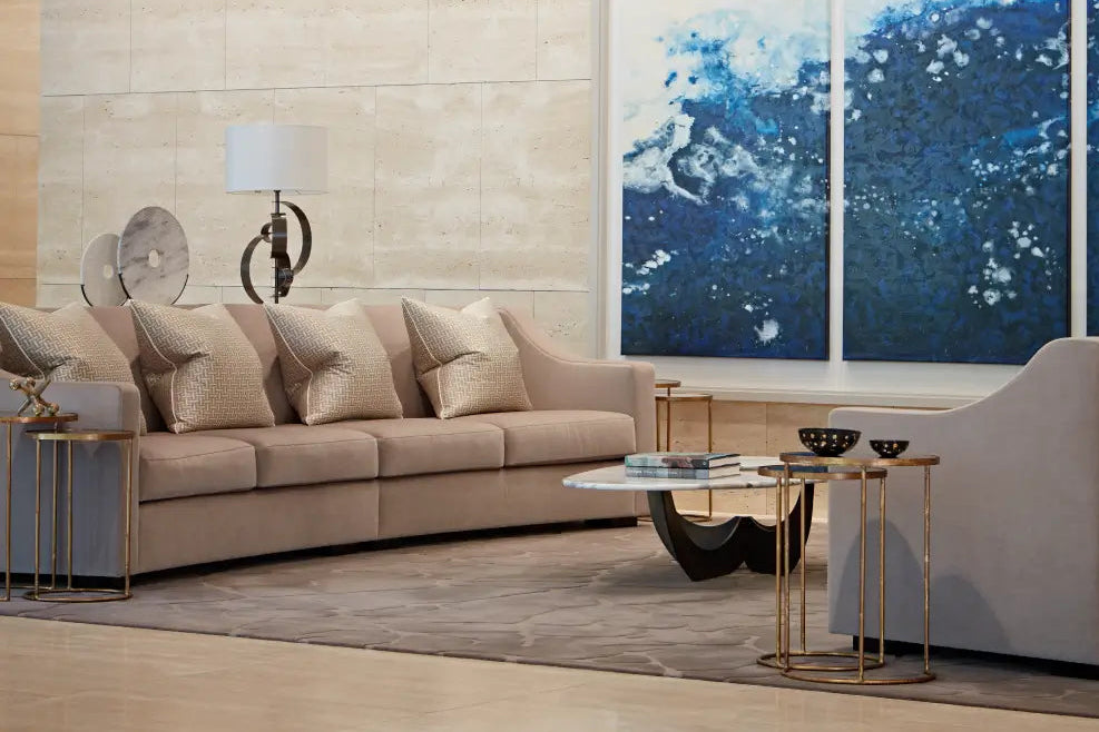 A modern living room featuring a beige sectional sofa with textured throw pillows, a marble-top coffee table with gold metal legs, and a large blue abstract wall art piece on a light stone wall.