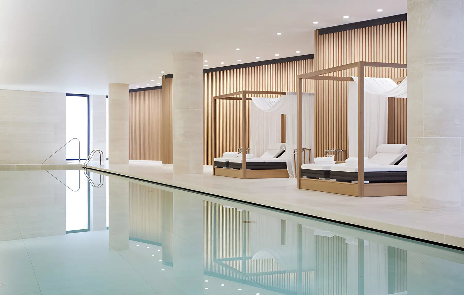 Luxury indoor poolside lounge area with two wooden canopy daybeds on a light stone terrace, reflecting in the calm water.