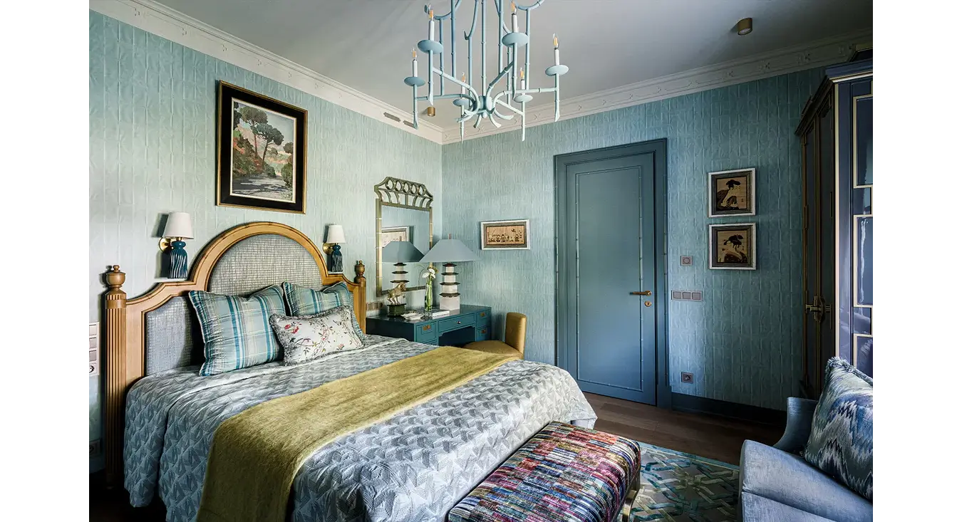 A styled bedroom interior with a bed dressed in blue patterned bedding and a yellow-green throw, set against light blue wallpapered walls. A chandelier hangs above, with framed wall art, bedside tables, and a doorway visible.