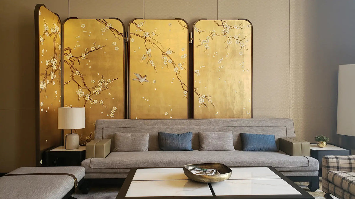 A luxury modern living room featuring a large four-panel gold folding screen with delicate white cherry blossoms and birds, positioned behind a light gray fabric sofa with blue accent pillows.