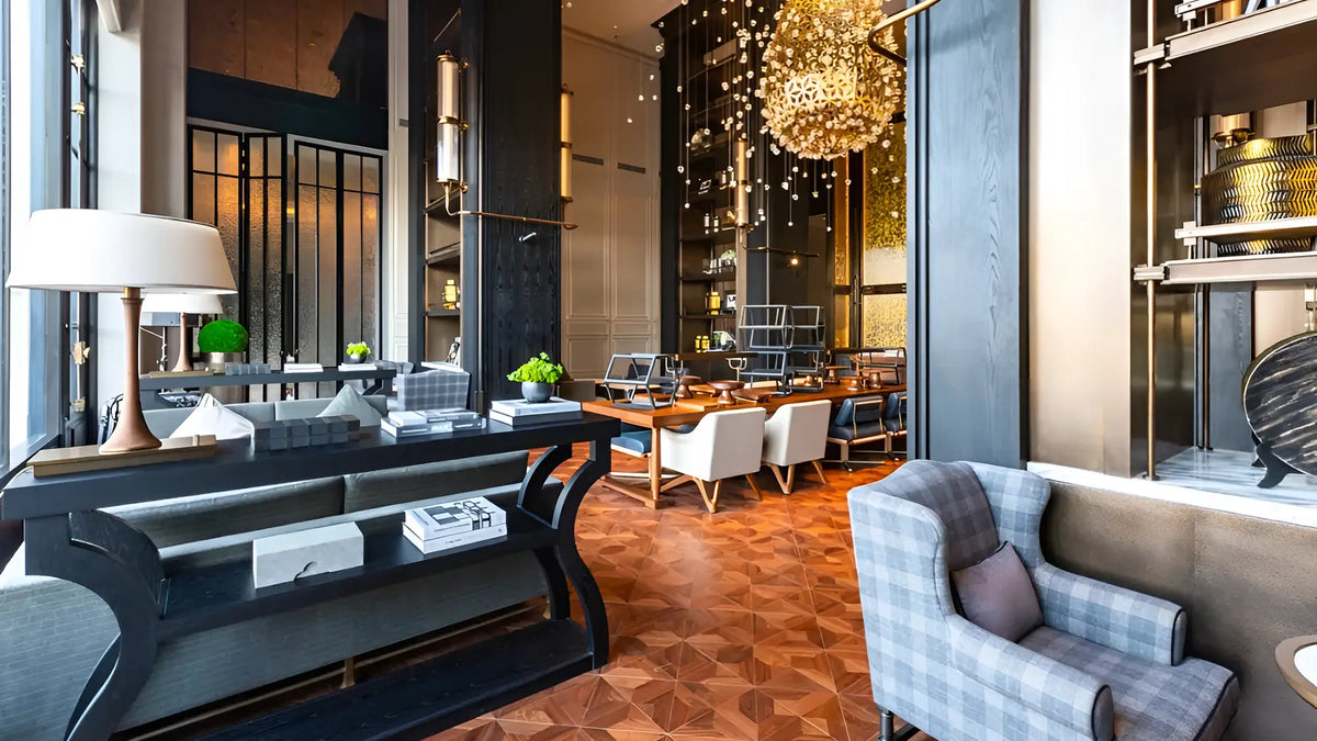 A luxury modern interior space featuring a stylishly decorated lounge area with a plaid armchair, a black console table, and a warm wood geometric patterned floor. In the background, a dining area with white chairs and a glowing crystal chandelier is visible.