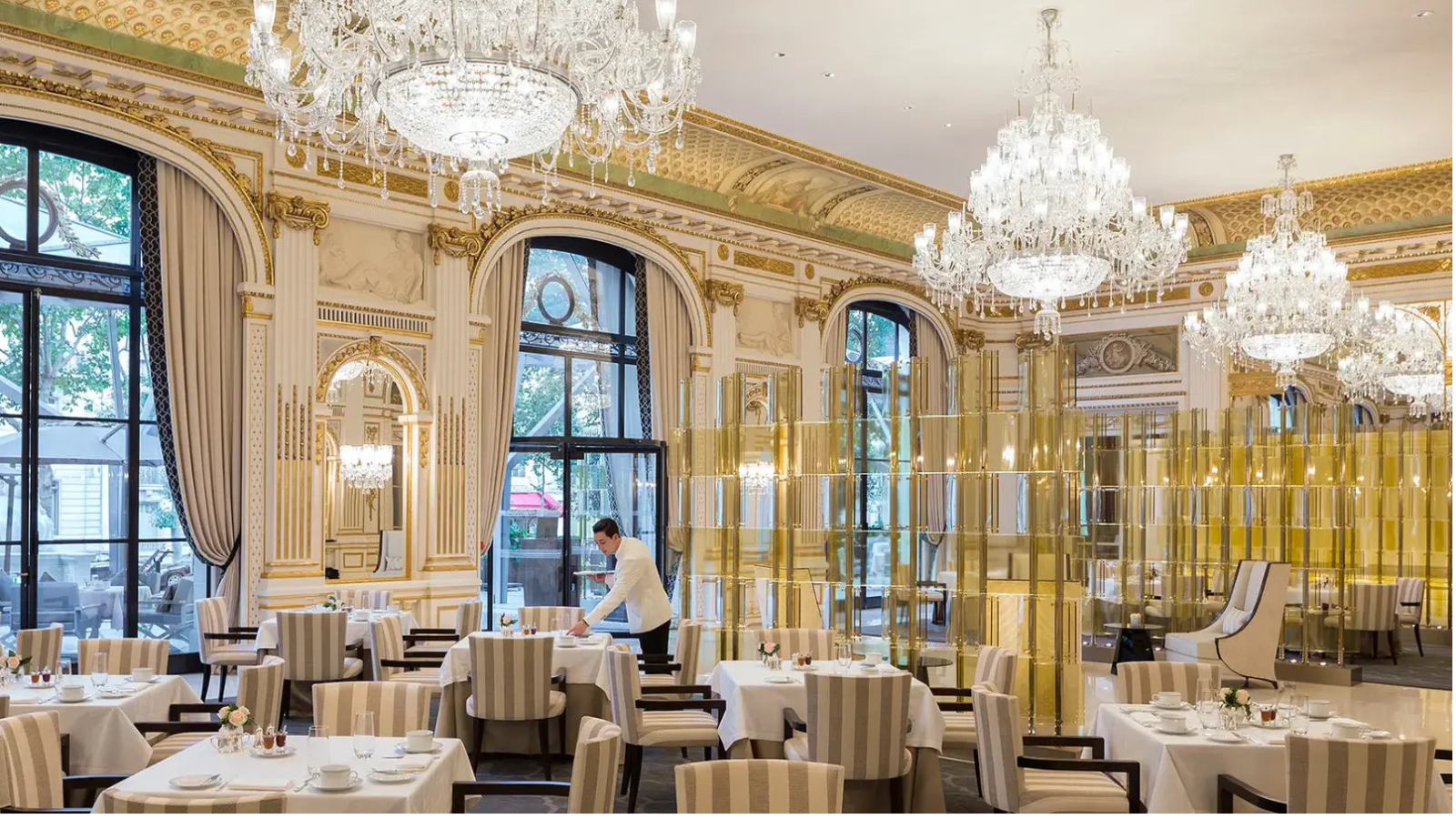 An elegant dining room with multiple large crystal chandeliers, cream and gold walls, arched windows with drapes, and many table settings with striped chairs. A waiter is visible near the center.