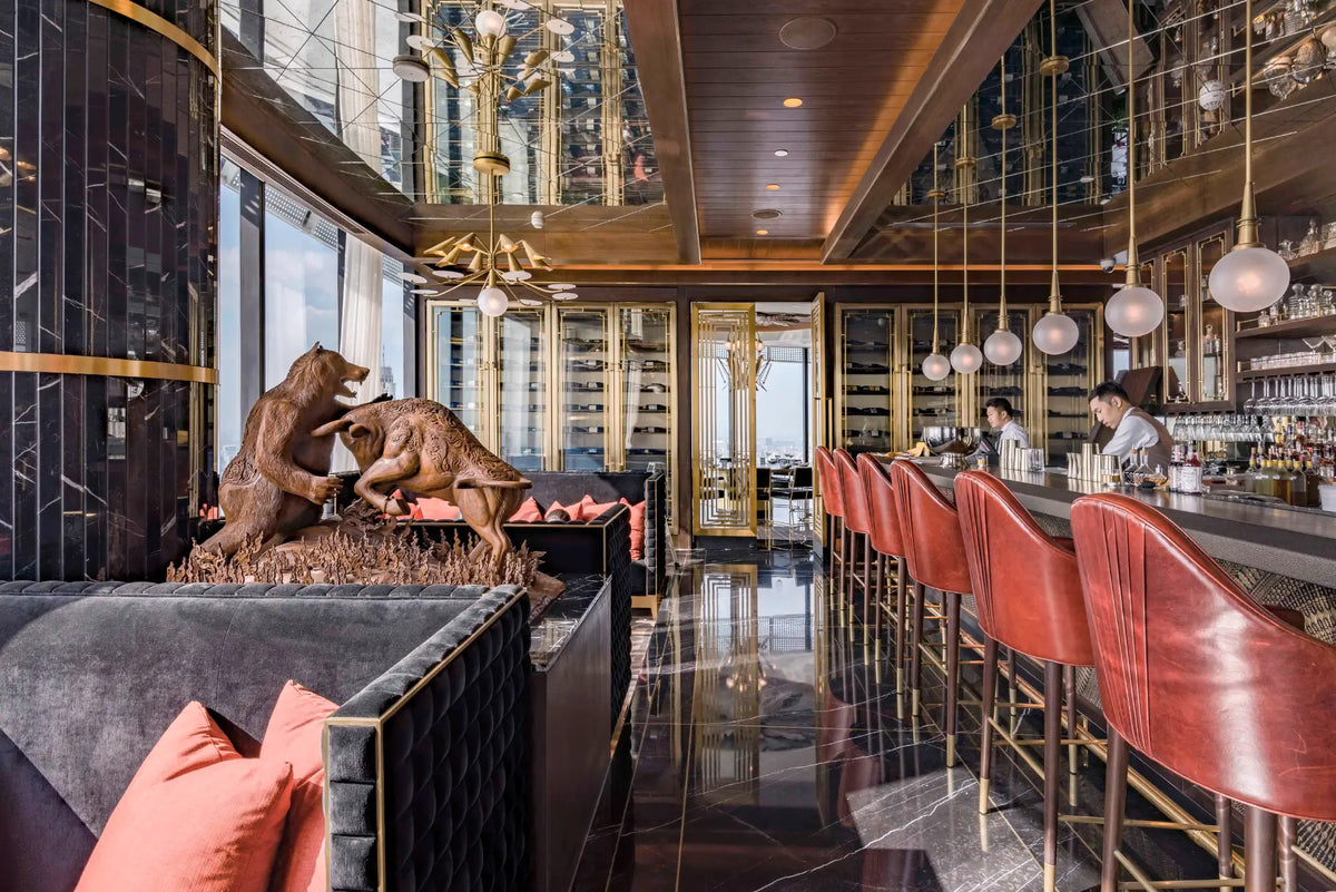 An upscale interior bar and lounge with a long row of red leather bar stools, dark marble flooring, mirrored walls, hanging pendant lights, and a sculptural bronze bear statue in the foreground.