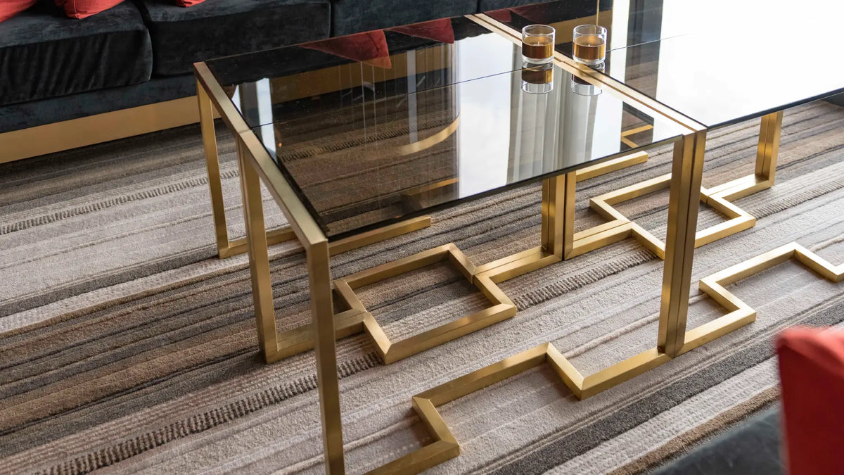A modern rectangular coffee table with a black glass top and geometric gold metal legs, styled in a contemporary living room setting on a striped rug.