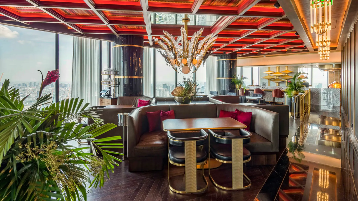 A luxury indoor lounge or restaurant interior with a red geometric ceiling, large chandeliers, upholstered booth seating with red pillows, bar stools, polished wood floors, and abundant green plants. Floor-to-ceiling windows show an outdoor city view.