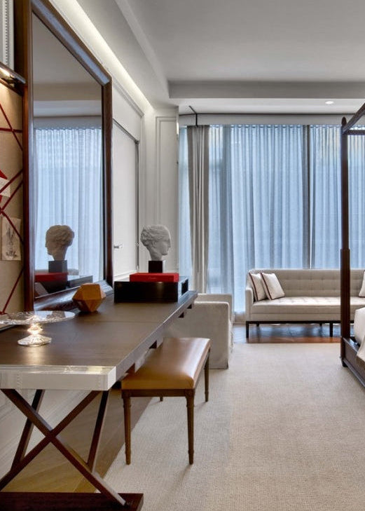 A modern bedroom interior featuring a wooden vanity table with a matching stool, a large wall mirror, and a four-post bed frame in the background near sheer curtains and a light beige sofa.