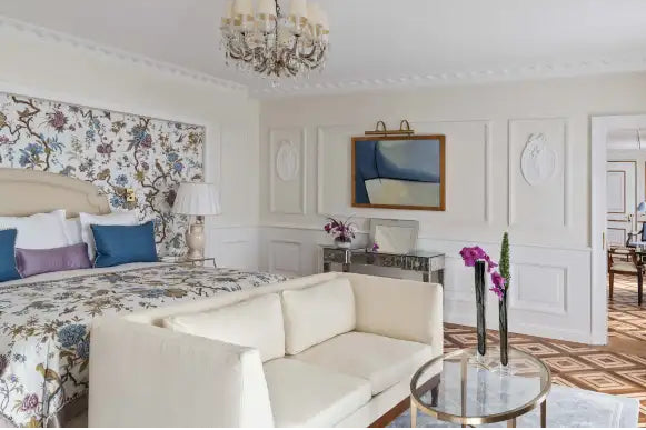 A bright, elegant bedroom interior featuring a bed with a floral wallpaper headboard, cream-colored furniture, a chandelier, and a patterned tile floor.