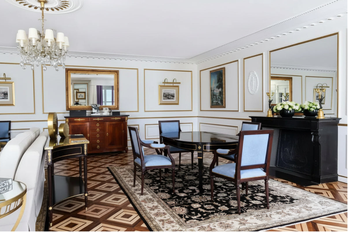 An elegant interior dining room with a dark wood dining table and four light blue upholstered chairs on a patterned area rug. The room features cream walls with gold trim, a large crystal chandelier, a wooden sideboard with mirrors, and a black console table with a floral arrangement.