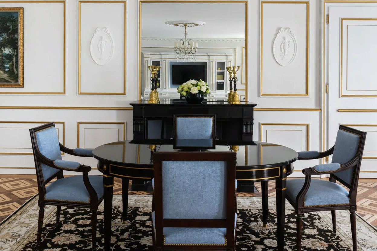 An elegant dining room setup featuring a round dark wood table with a black marble top, surrounded by four matching armchairs with light blue upholstery. The room is decorated with white walls, gold trim, a large wall mirror, and a patterned rug.
