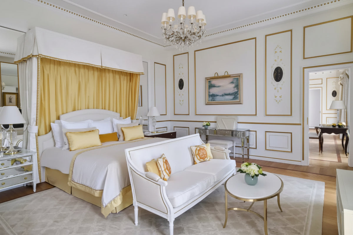 A luxurious bedroom interior featuring a white four-poster bed with gold drapery, a white upholstered bench with gold-trimmed pillows, a round white coffee table with gold legs, and elegant cream and gold paneled walls.