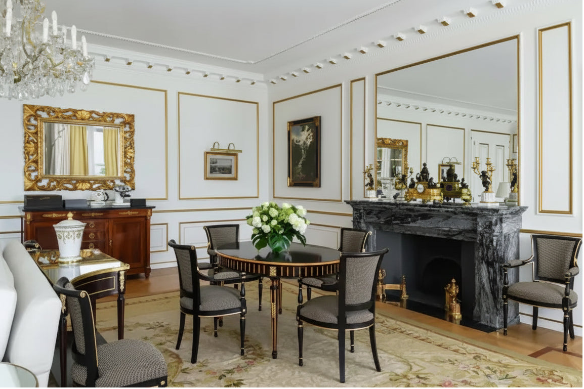 An elegant interior room featuring a dark wood dining table with six upholstered chairs, a large crystal chandelier, a white sofa, a dark marble fireplace with gold detailing, and ornate gold-framed mirrors on white paneled walls.