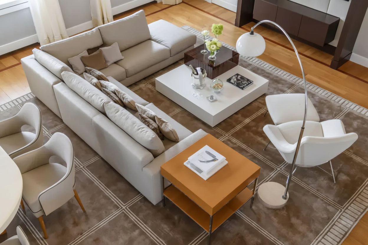 A modern living room setup featuring a large light beige sectional sofa, a white coffee table, a wooden side table, two white accent chairs, a tall floor lamp, and a patterned brown area rug on a wood floor.