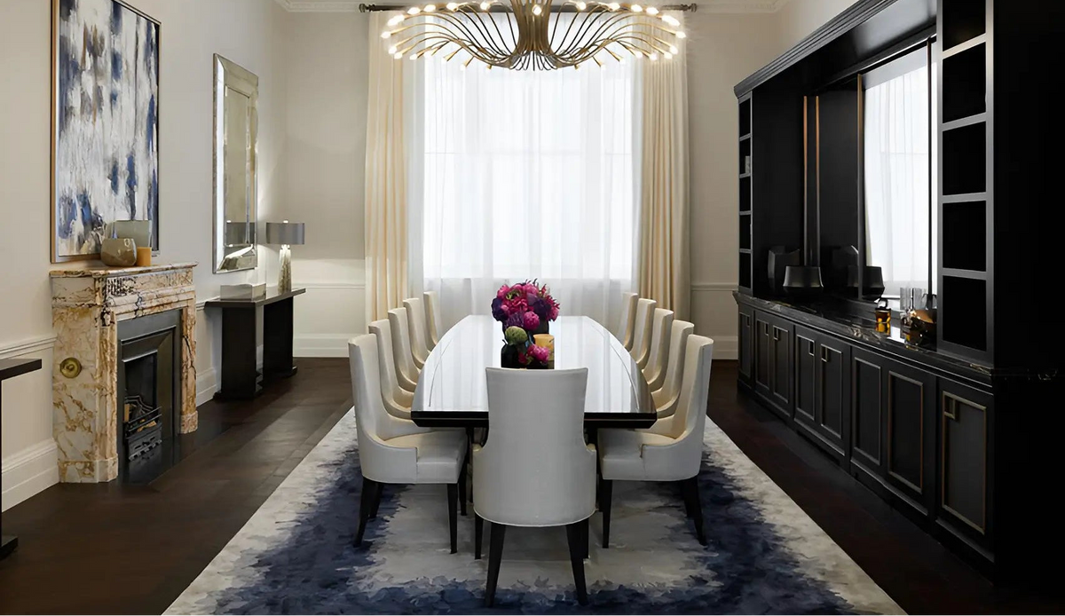 A luxury dining room featuring a long white table, white upholstered chairs, a large blue and white area rug, a black wall-mounted cabinet, and a decorative chandelier.