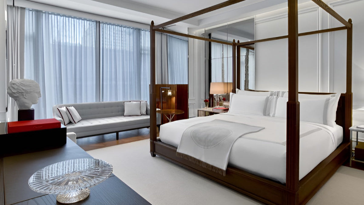 A luxury bedroom featuring a dark wood four-poster canopy bed with crisp white bedding, set against a white paneled wall with a large window and sheer curtains.