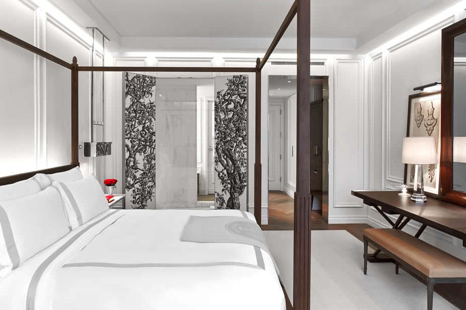 A luxury four-poster bed in a modern, elegant bedroom with white linens, dark wood frame, a vanity desk with a mirror, and a bench.