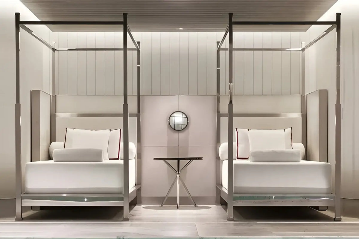A luxury bedroom interior featuring two matching white beds with slim silver metal four-post canopies, a small central side table, and a round wall mirror against a light wood-paneled background.