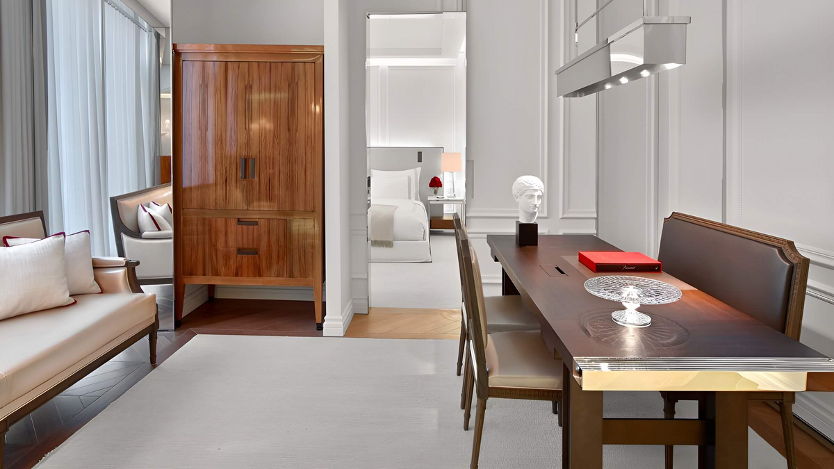 A modern dining room scene featuring a dark wood dining table with gold-toned metal trim, matching wooden chairs, a beige upholstered bench, and a tall wooden cabinet in a bright, contemporary interior.