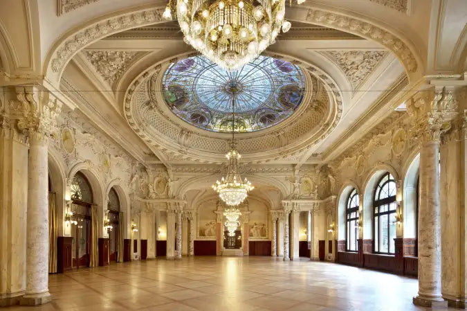 An ornate grand hall with a large central stained-glass dome, multiple crystal chandeliers, arched windows, and decorative columns. The interior features cream-colored plasterwork, marble-like columns, and a polished floor.