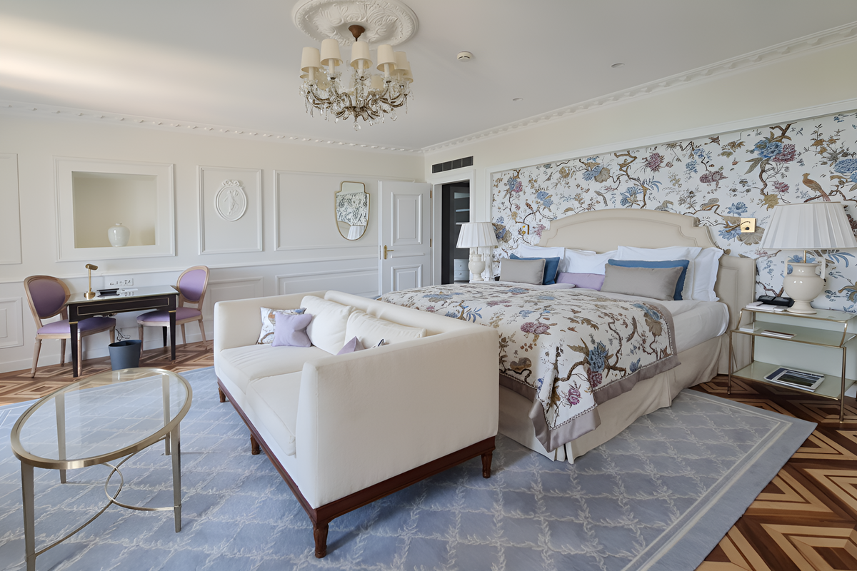 A luxurious bedroom interior featuring a large bed with a floral bird-patterned quilt and matching wallpaper, a cream upholstered armchair, a blue patterned area rug, and a dressing table setup in the background.