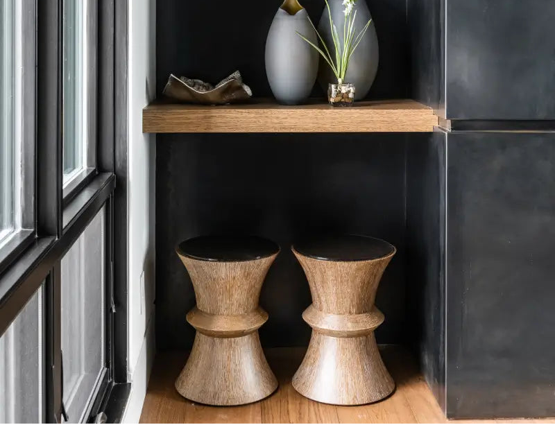 Two wooden pedestal stools in a modern interior setting with a dark paneled wall, wooden floor, and a floating shelf holding decorative vases.