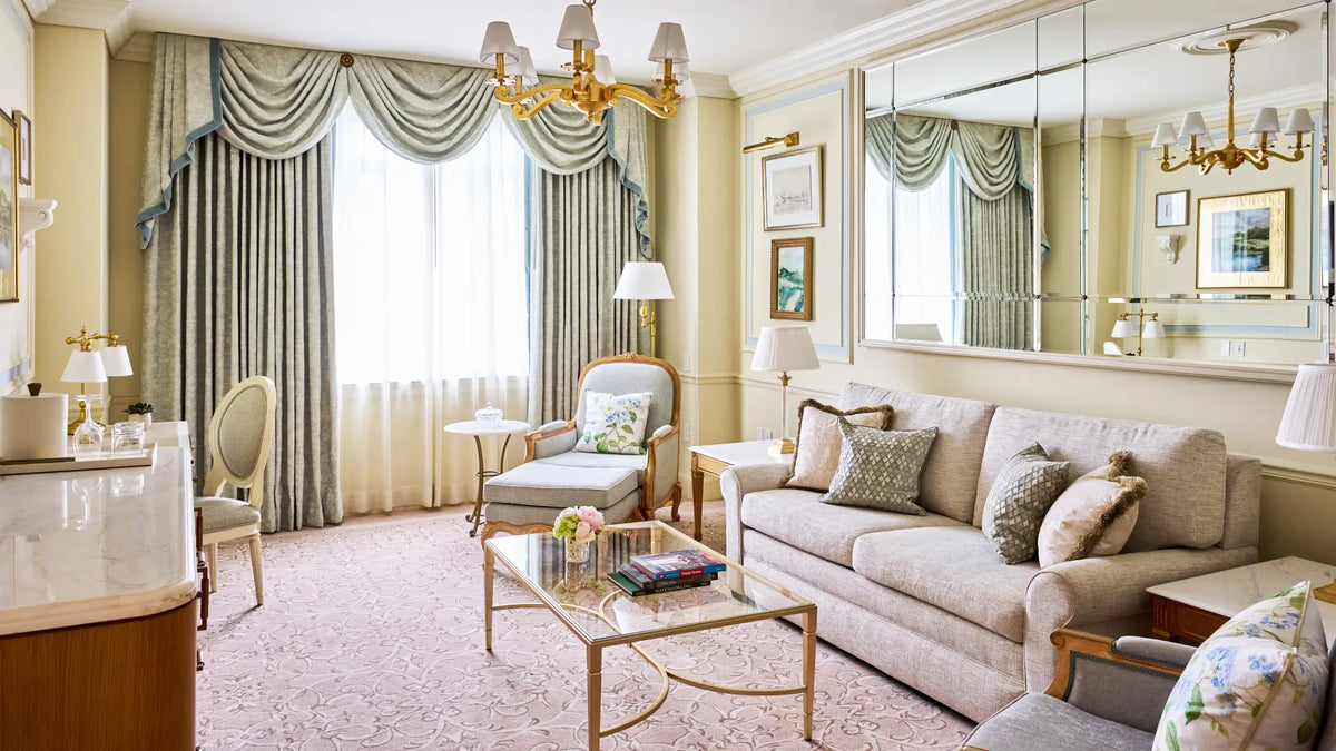 A luxurious living room featuring a light beige sofa, a matching armchair, a glass-top coffee table with gold accents, and elegant draped sage green curtains with gold-trimmed valances.