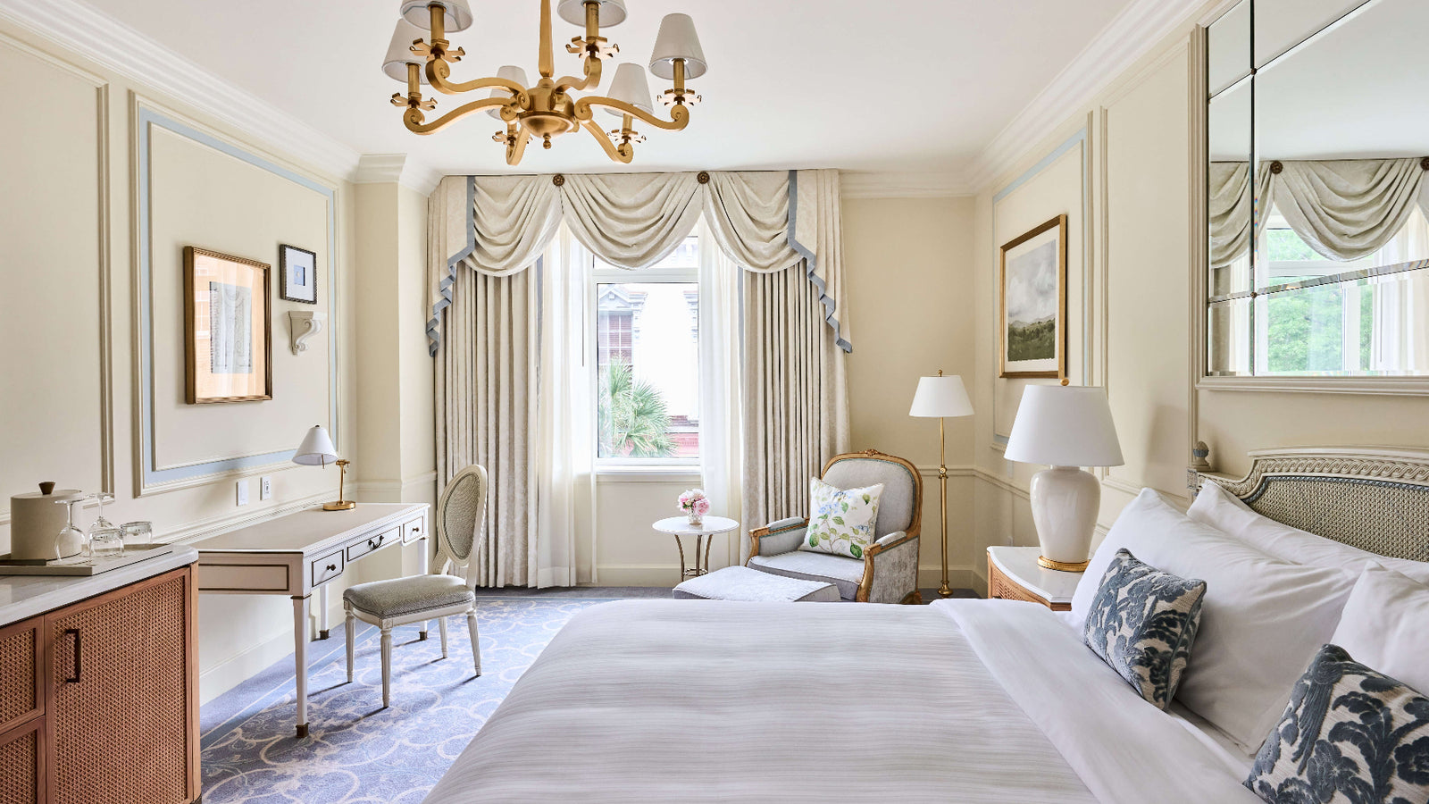 A luxurious hotel bedroom featuring a large bed with white linens and patterned blue pillows, a gold chandelier, a vanity desk, and elegant cream drapes by a bright window.