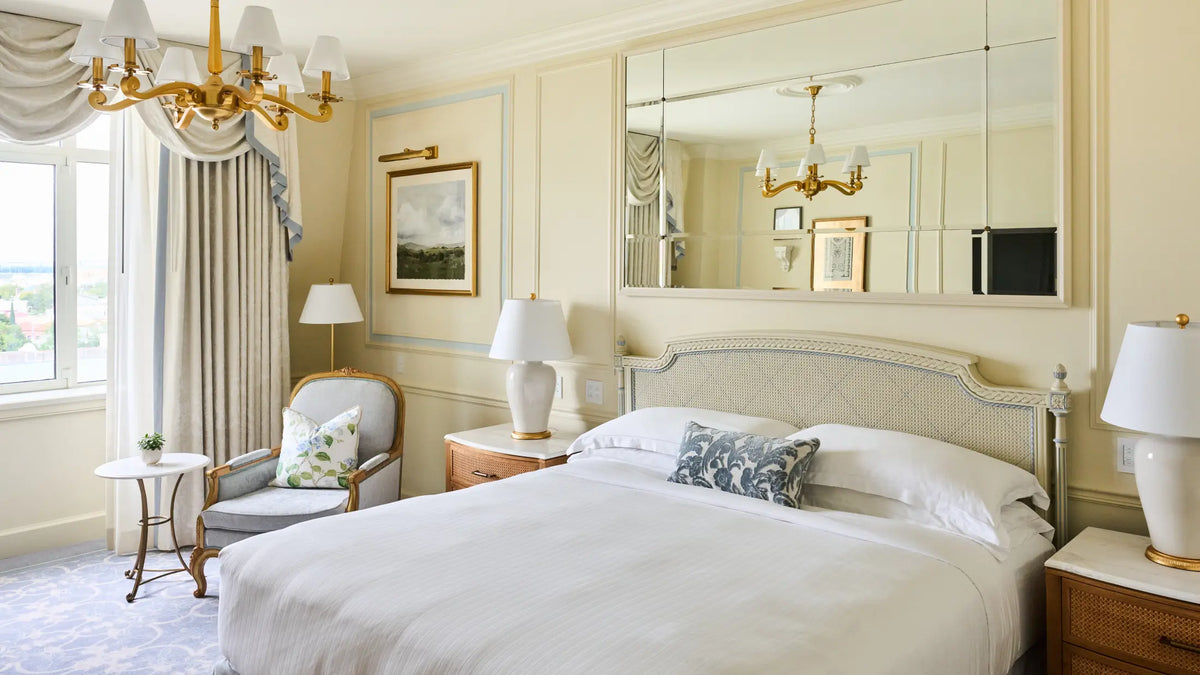 A luxurious bedroom featuring a large bed with white linens, a decorative blue and white pillow, a large wall mirror, a gold chandelier, and elegant cream-colored walls.