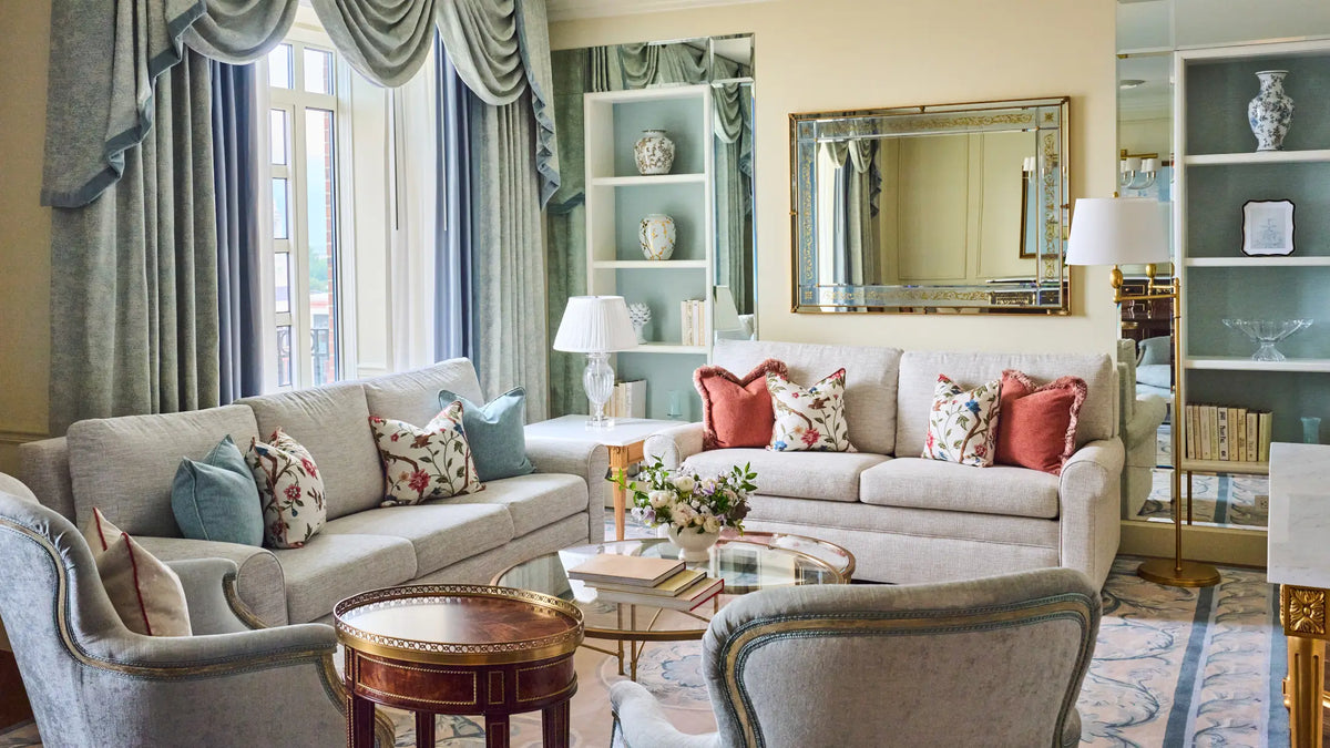 A styled living room featuring a light beige sofa with decorative throw pillows, a matching armchair, a round wooden coffee table, and a large rectangular mirror on a cream wall.