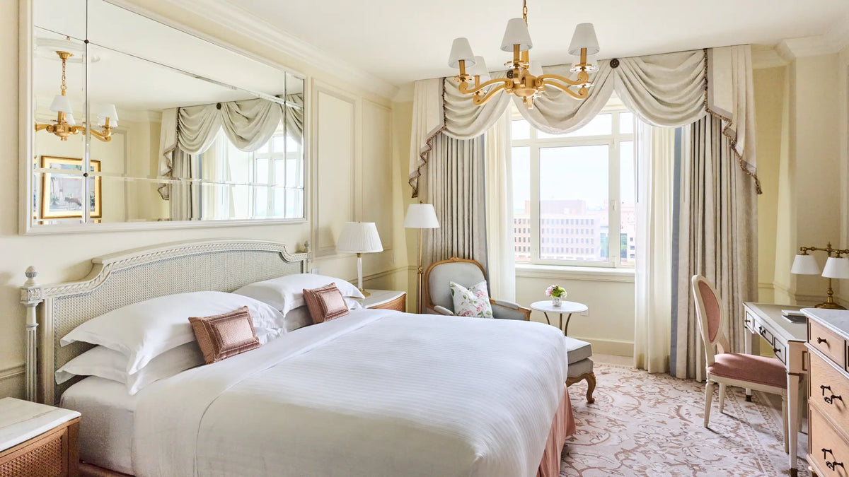 A luxurious bedroom interior featuring a large bed with white linens and pinkish-brown accent pillows, a gold chandelier, a large wall mirror, and a window with elegant drapes and sheer curtains.