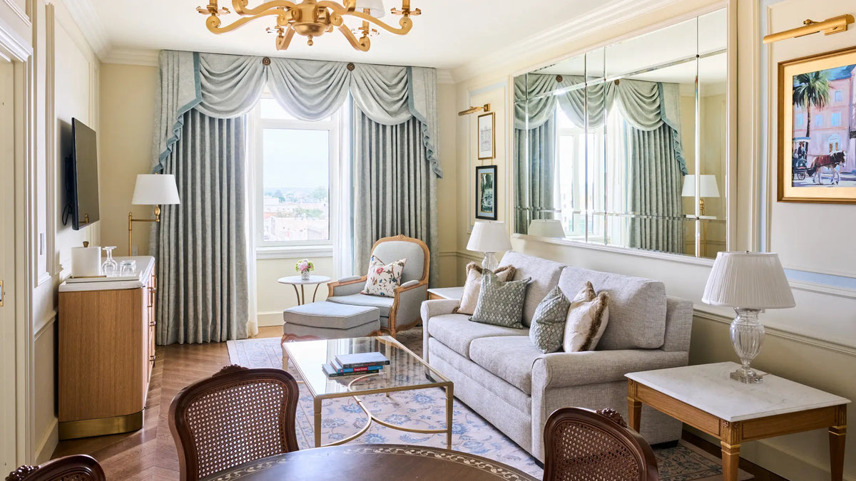 A luxury hotel suite living room featuring a light beige sofa, matching armchair, blue and gray striped curtains, a glass coffee table, and a large wall mirror reflecting the window view.