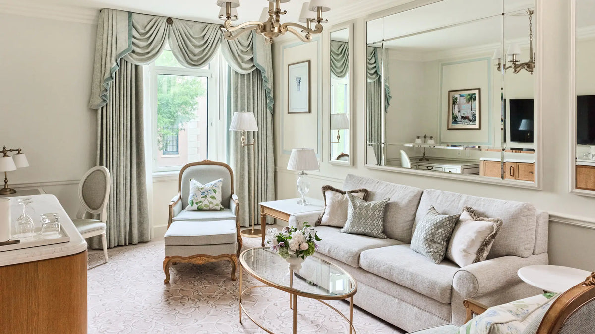 A luxury living room featuring a light beige sofa, a matching armchair, elegant draped curtains, a large wall mirror, and a glass-top coffee table.
