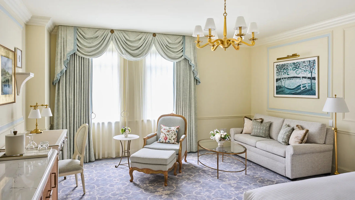 A luxury hotel-style living room featuring a light gray sofa, a matching armchair and ottoman, a gold chandelier, and elegant drapes in sage green and blue-gray tones.