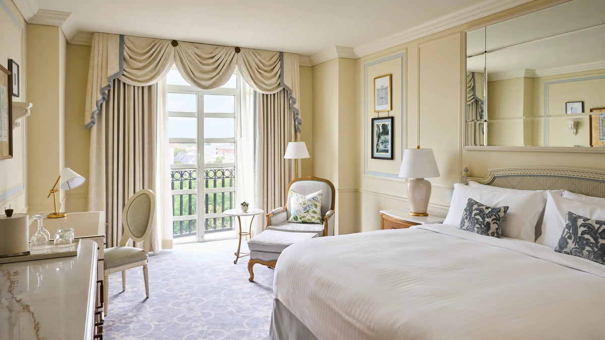 A luxurious hotel bedroom featuring a large bed with white linens and patterned pillows, cream walls, draped window curtains, a matching armchair, and a vanity desk.