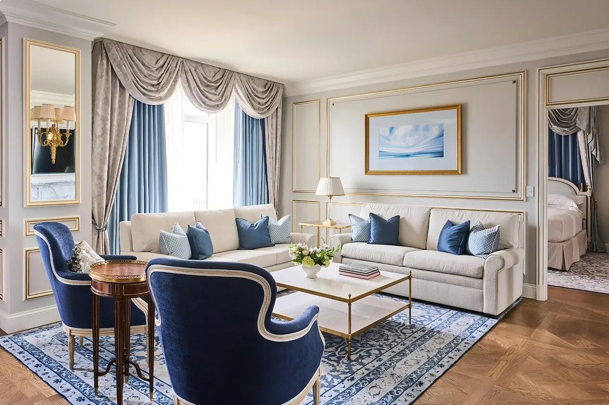 A luxury living room featuring a cream sectional sofa with blue accent pillows, two navy blue velvet armchairs, a patterned blue and white rug, and elegant gold-trimmed wall molding.