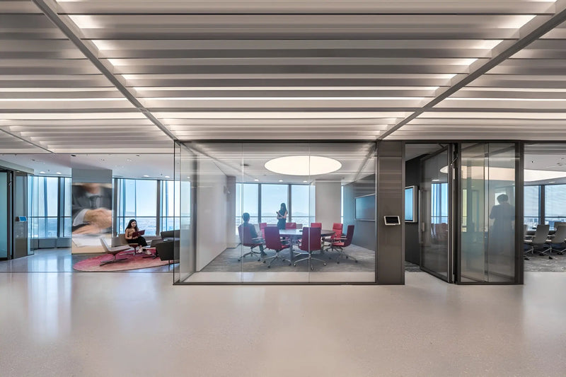 A modern glass-walled office meeting room with a round ceiling light, surrounded by a large open floor space with a slatted ceiling and office seating.