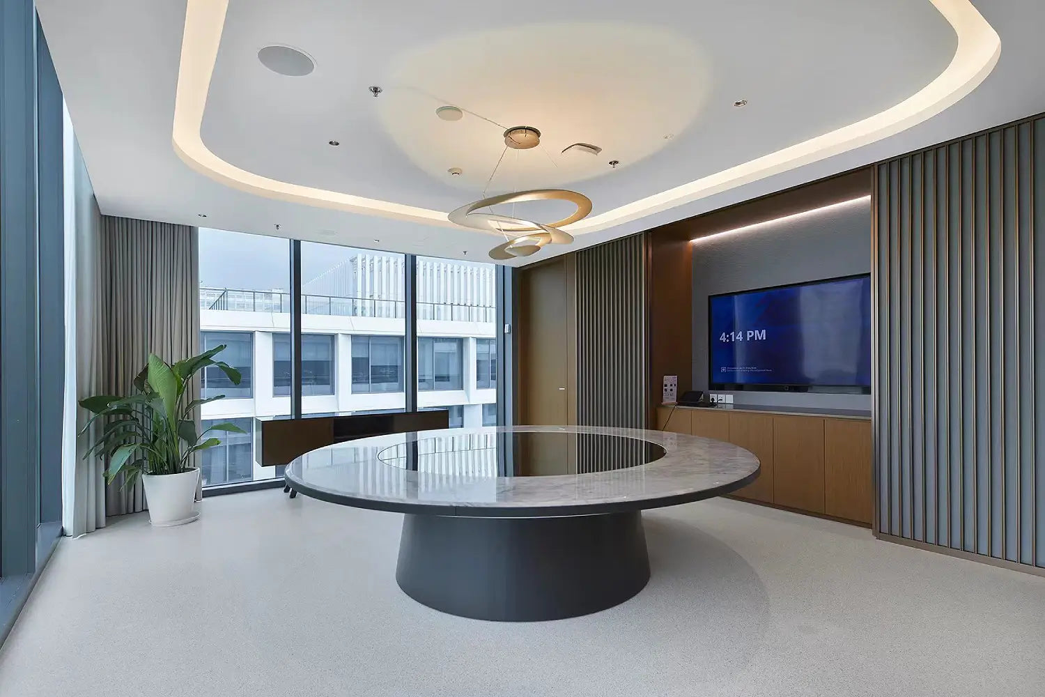 A modern conference room featuring a large oval marble-top table with a dark circular base, a wall-mounted television, and contemporary ceiling lighting.