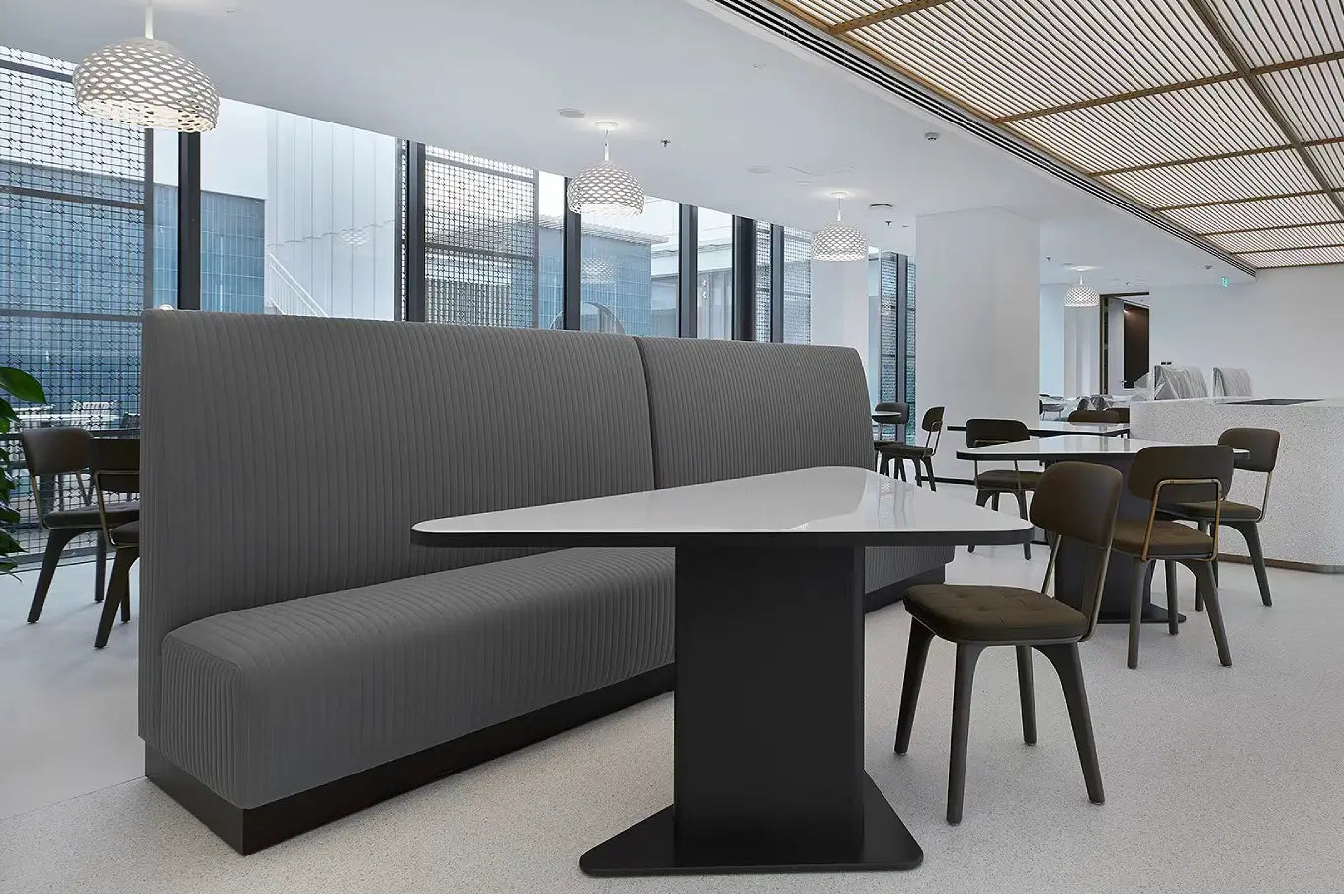 Modern dining area with a gray ribbed upholstered booth, white oval table, and dark wood chairs in a bright interior with large windows and a slatted ceiling.