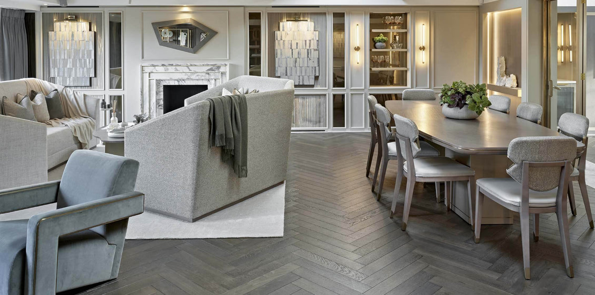A luxury modern living and dining room interior featuring a light gray upholstered dining set, a gray armchair, a white sofa, herringbone wood flooring, and elegant built-in shelving.