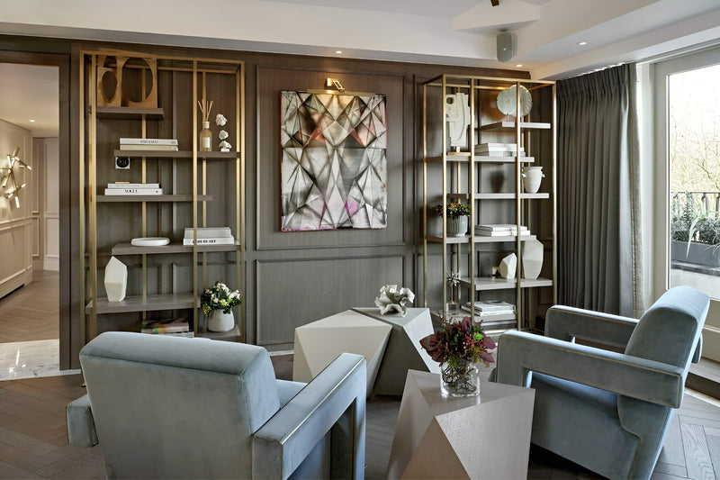 A modern living room featuring two light blue-gray velvet armchairs, geometric white side tables, and a large abstract geometric wall art piece hanging in the center. The background consists of dark gray wood paneling with gold-framed shelving units containing decorative objects.